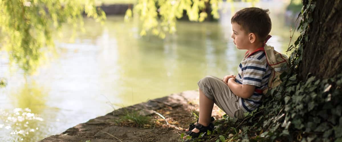 A young boy sits against a tree trunk and looks over a pond